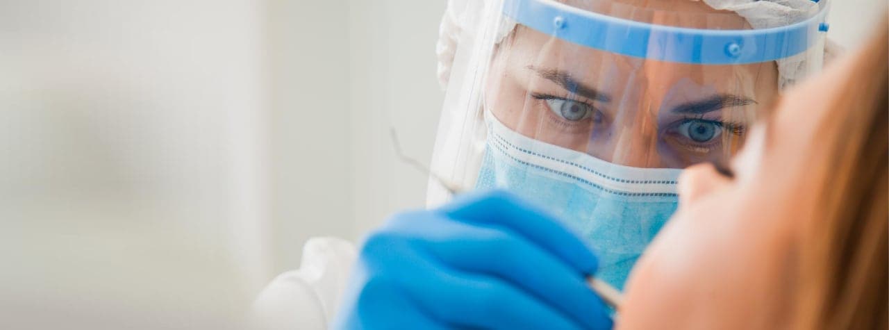 A dental professional wearing safety PPE gear and white lab coat smiling compassionately